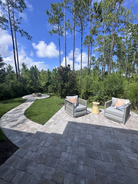 A serene patio with cozy wicker chairs, a small table, and a pathway leading to a fire pit, surrounded by lush greenery.