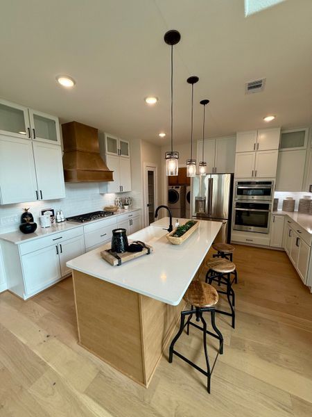 A modern kitchen with a white island, pendant lighting, and wooden stools, featuring sleek cabinetry and stainless appliances.
