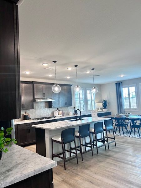 A modern kitchen featuring dark cabinets, a marble island, sleek pendant lights, and a cozy dining area with large windows.