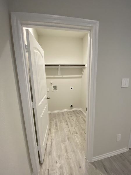 A compact utility room with light wood flooring, white walls, and a shelf for storage.