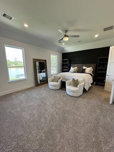 A cozy bedroom featuring plush carpeting, elegant seating, a large mirror, and scenic lake views through ample windows.