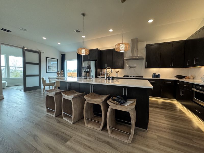 A modern kitchen with sleek dark cabinets, island seating, pendant lighting, and a bright dining area.