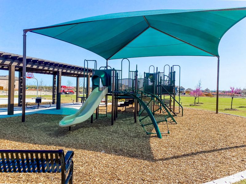 Community playground with teal shade canopy, slide, and wood chip ground cover near a pergola and basketball hoop Community playground with teal shade canopy, slide, and wood chip ground cover near a pergola and basketball hoop