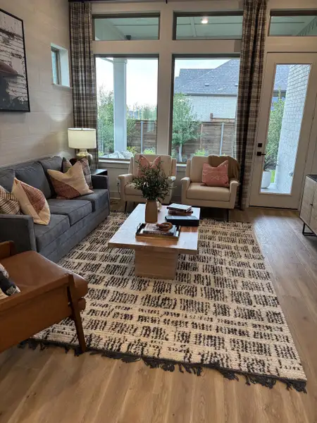 A cozy living room featuring a large patterned rug, comfy seating, and abundant natural light through tall windows.
