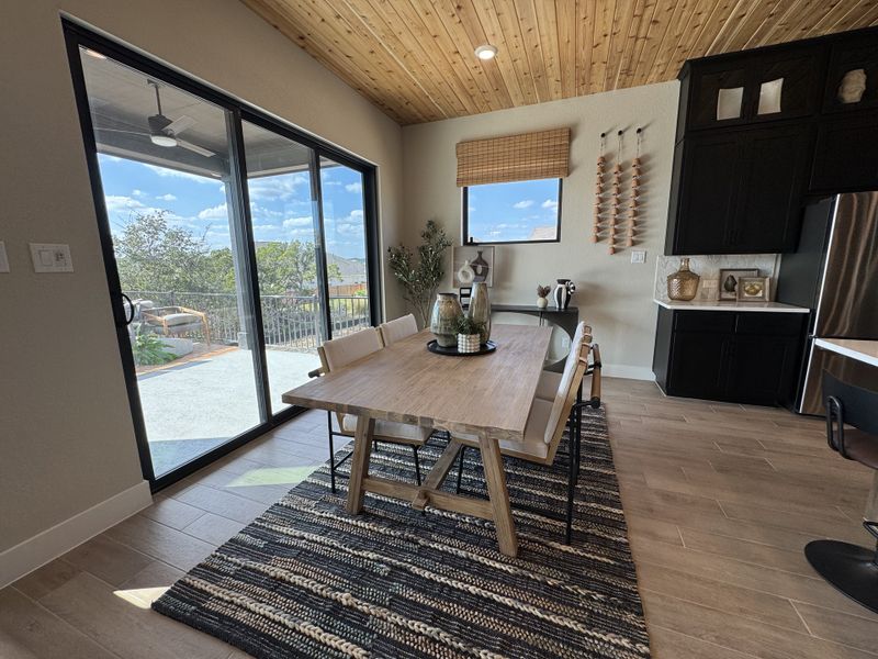 A dining area with a wooden table, wicker accents, and large glass doors opening to a patio, set beneath a wooden ceiling.