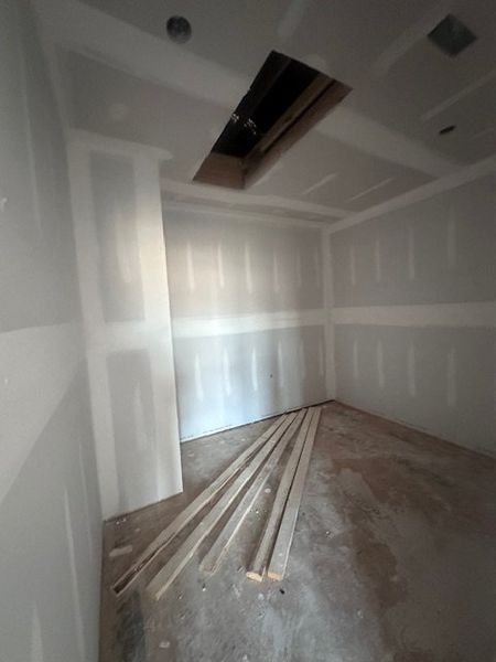 An unfinished room with drywall, an open ceiling section, and scattered wooden planks awaiting completion.