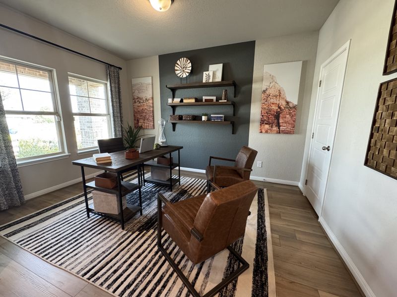 A modern home office with a wooden desk, leather chairs, wall art, and natural light from large windows.