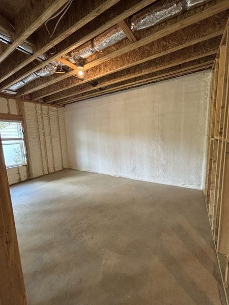 A spacious unfinished room with spray foam insulation, exposed beams, and natural light from a window.
