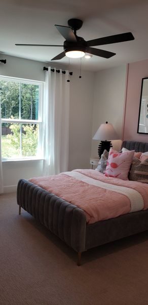 A cozy bedroom with a modern ceiling fan, plush bed, soft pink accents, and a large window with sheer curtains.