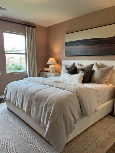 A cozy bedroom with a plush bed, soft beige hues, elegant artwork, and natural light filtering through a window.