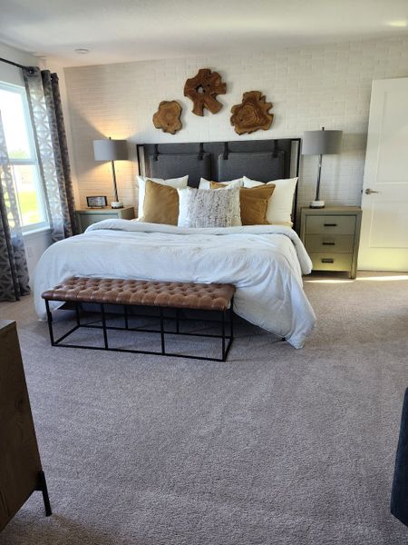 A cozy bedroom with a plush bed, decorative pillows, and wood wall art, accented by natural light and soft carpeting.