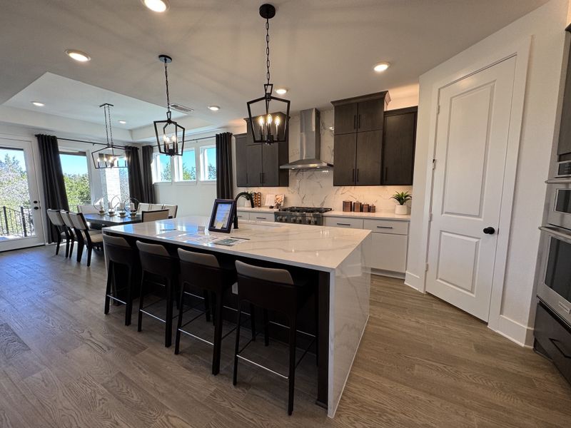 A spacious kitchen with dark cabinets, marble island, modern lighting, and a dining area with natural light from large windows.