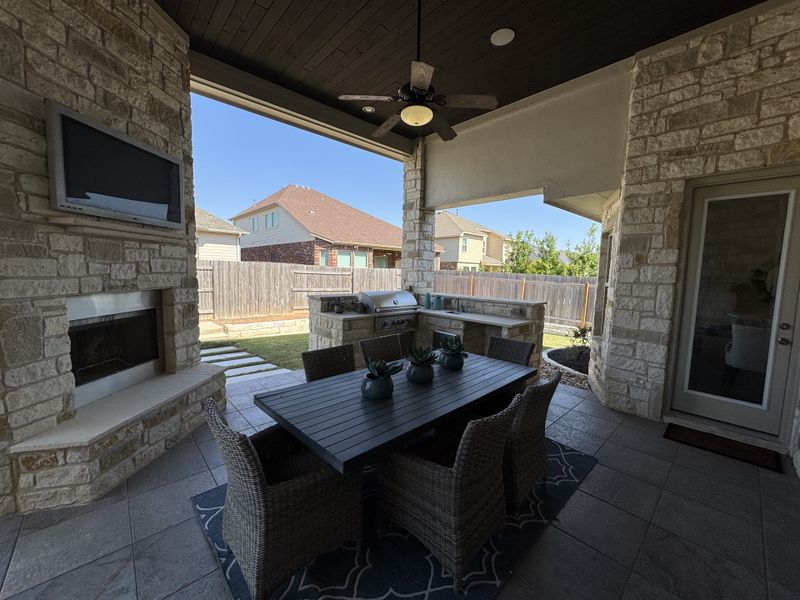 A cozy outdoor patio with a stone fireplace, TV, dining set, and built-in grill, perfect for entertaining.