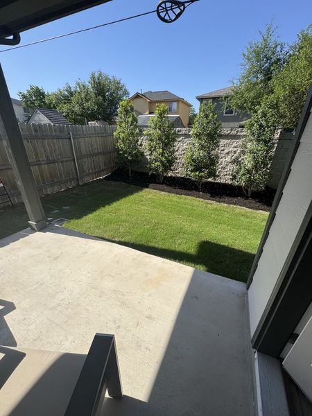 A cozy backyard patio with lush grass, wooden fencing, and young trees under a clear blue sky.