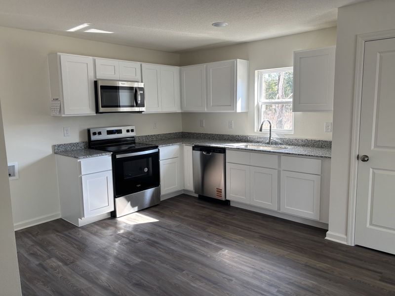 A modern kitchen with white cabinets, granite countertops, and stainless steel appliances, featuring wood-style flooring.