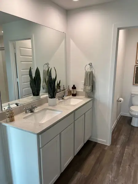A modern bathroom features dual sinks, sleek countertops, and elegant wood flooring, accented by vibrant greenery.