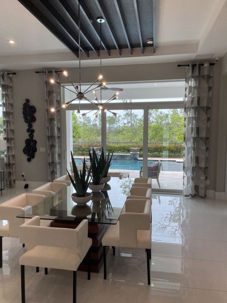 A sleek dining area with a glass table, modern chandelier, and pool view, featuring elegant white chairs and greenery accents.