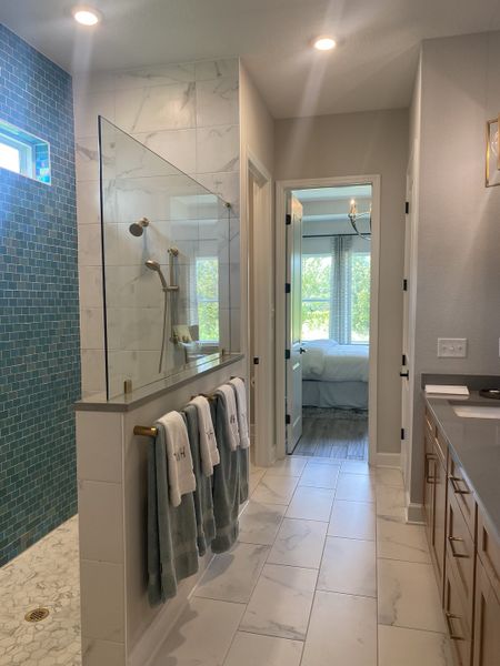 An elegant bathroom with a glass-enclosed shower, marble tiles, and wood cabinetry, leading into a sunlit bedroom.