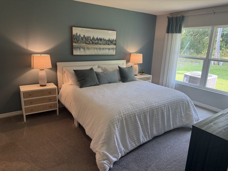 A cozy bedroom featuring a soft blue accent wall, plush carpeting, and a large window with a scenic backyard view.