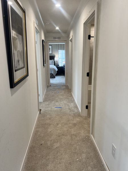A cozy hallway with carpeted floors, recessed lighting, and framed art leading to a bright bedroom.