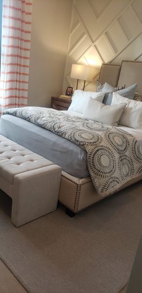 A cozy bedroom with a patterned duvet, plush beige bench, and accent wall with geometric paneling.