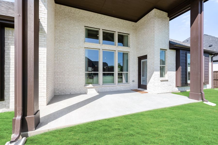 Covered back patio with concrete slab, white brick exterior, large windows, and green lawn