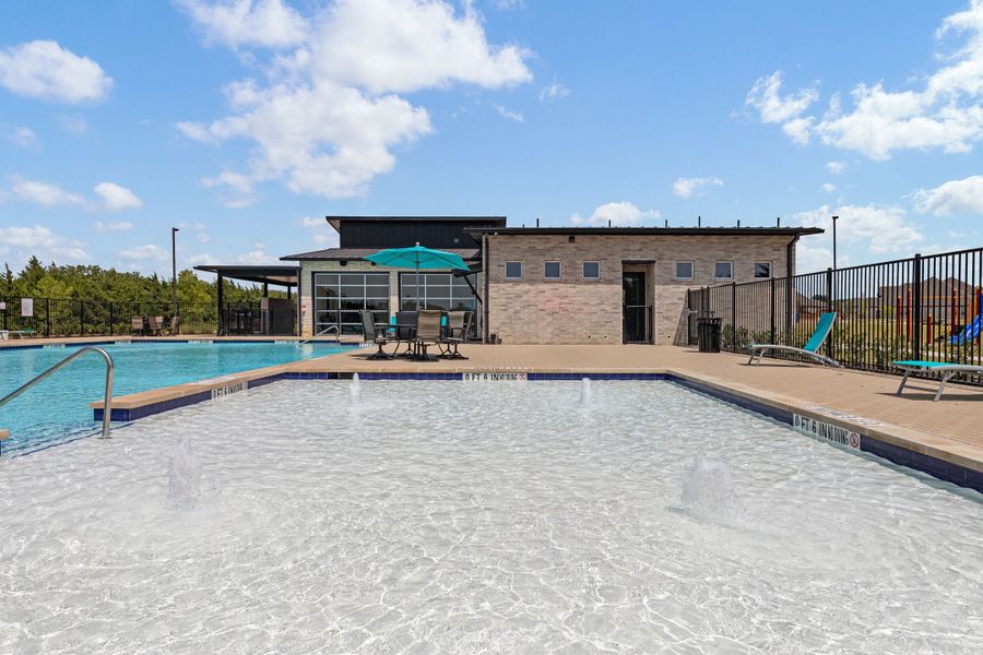 Community pool with splash pad, lounge chairs, and brick clubhouse building behind black iron fence
