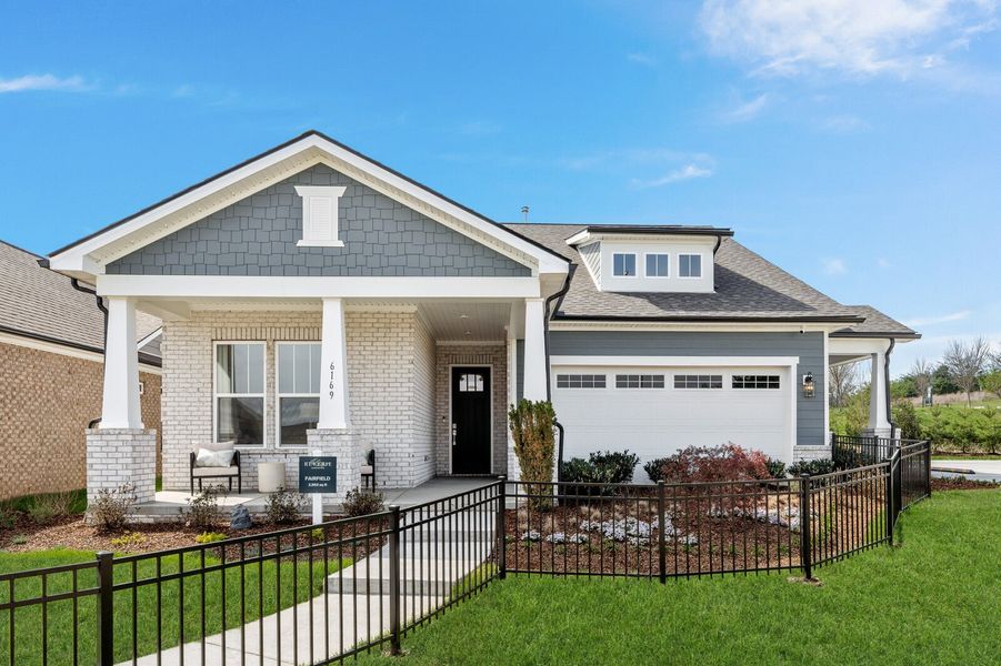Single-story craftsman home with gray and brick exterior, covered porch, two-car garage, and black iron fence.