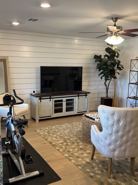 A cozy living room with shiplap walls, exercise bike, tufted chair, and a large TV on a rustic console.