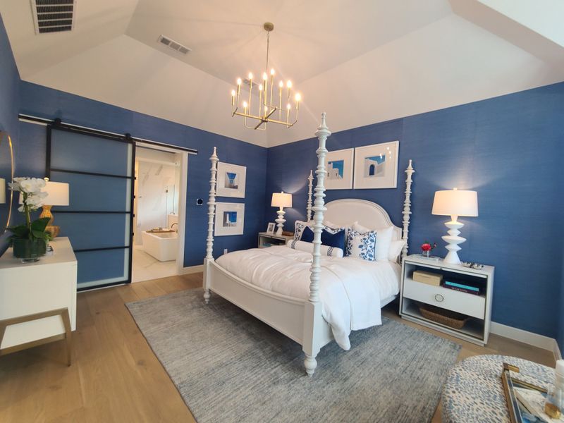 A chic bedroom with blue walls, white four-poster bed, elegant lighting, and a barn door leading to a stylish bathroom.