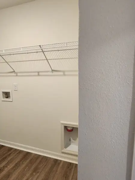 A functional laundry room with a hardwood floor, utility connections, and a wire shelving unit for storage.