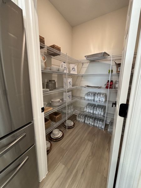 A well-organized pantry with wire shelving, stocked with essentials and dishes, featuring light wood floors and an open design.
