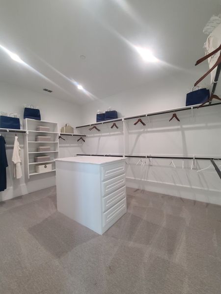 A spacious walk-in closet featuring white shelving, ample hanging space, and a central island with drawers. A spacious walk-in closet featuring white shelving, ample hanging space, and a central island with drawers.