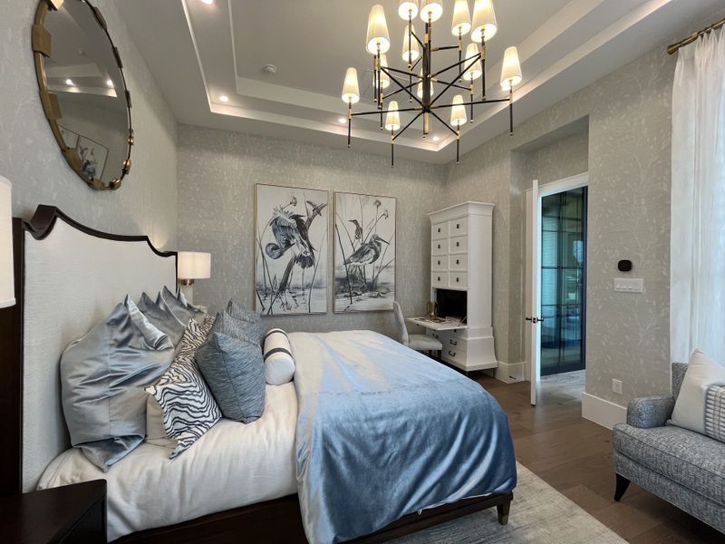 Elegant bedroom with modern chandelier, upholstered bed, and serene artwork, highlighted by soft grey tones and plush textures.