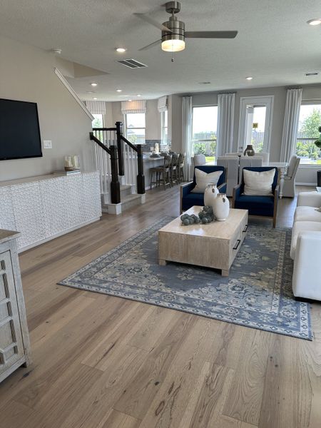 A spacious living area with light wood floors, modern furnishings, and natural light streaming through large windows.