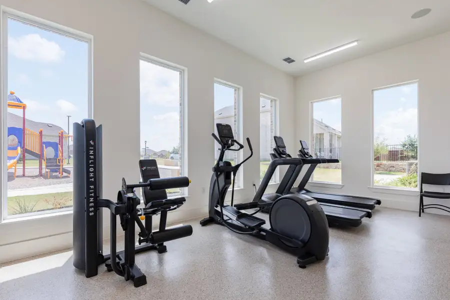 Community fitness room with elliptical, treadmill, and weight machine beside large windows overlooking a playground
