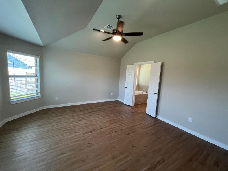 Spacious bedroom with wood flooring, neutral walls, and a ceiling fan. Open door reveals an adjoining bathroom.