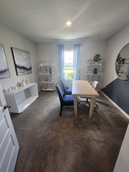 A cozy home office with a wooden desk, dark chairs, abstract art, and natural light filtering through soft curtains.