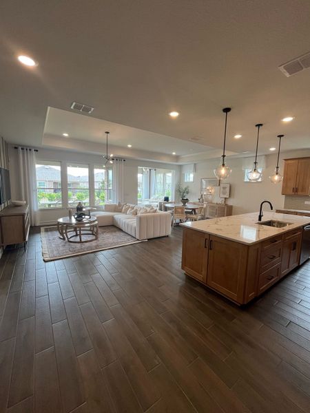An open-concept living area with wood floors, modern lighting, and large windows. Features a cozy sectional and kitchen island.