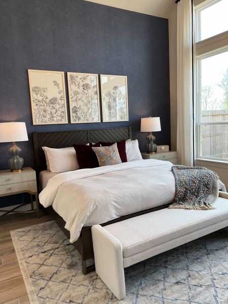 A cozy bedroom with a plush bed, elegant navy accent wall, floral art, and ample natural light through large windows.
