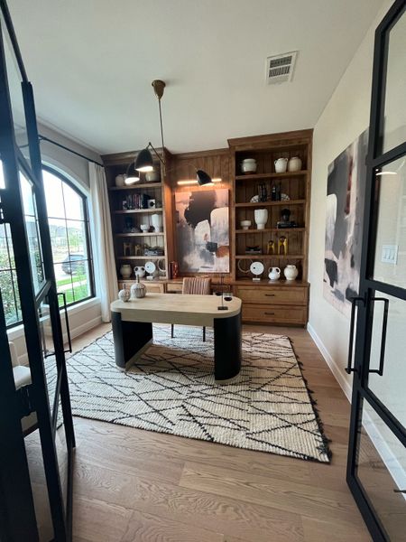 A stylish home office with wooden shelves, modern desk, abstract art, and large window with natural light.