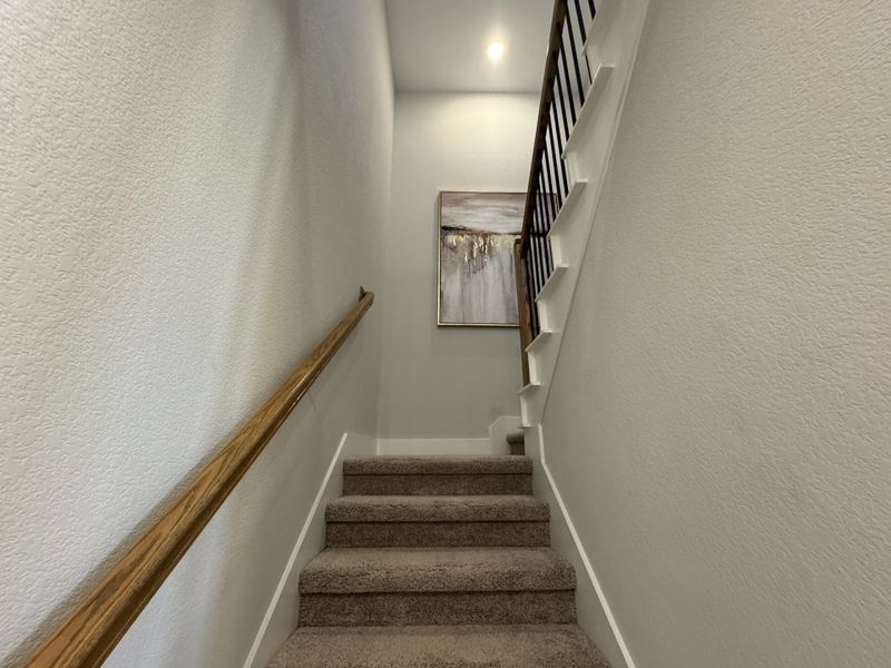 A carpeted staircase with a wooden handrail and elegant artwork on the wall, leading to a warmly lit upper floor.