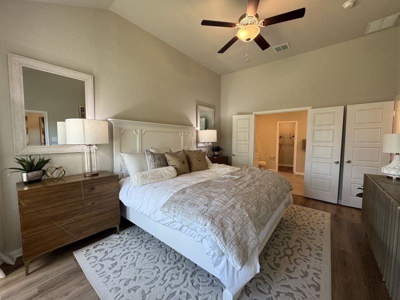 A cozy bedroom with a plush bed, wood dresser, elegant decor, and a ceiling fan on a high ceiling.