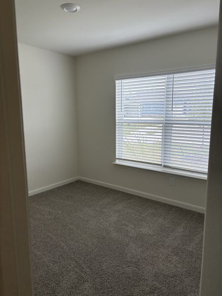 A cozy bedroom with plush carpeting, neutral walls, and ample natural light from a wide window with blinds.