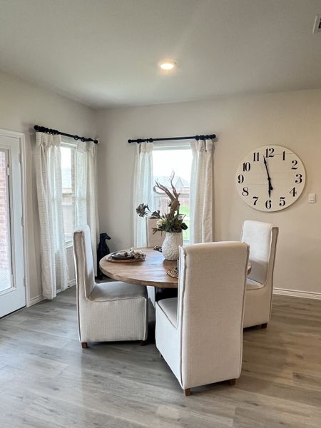 A cozy dining nook with a round wooden table, elegant beige chairs, large wall clock, and soft curtains letting in natural light. A cozy dining nook with a round wooden table, elegant beige chairs, large wall clock, and soft curtains letting in natural light.