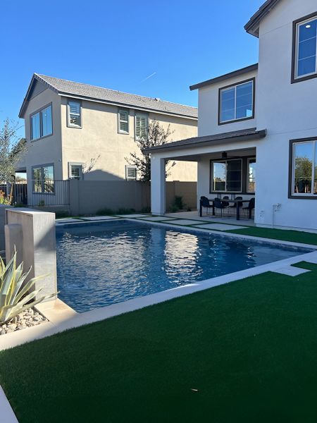  A luxurious backyard with a modern pool, artificial grass, and a covered patio area for outdoor dining.