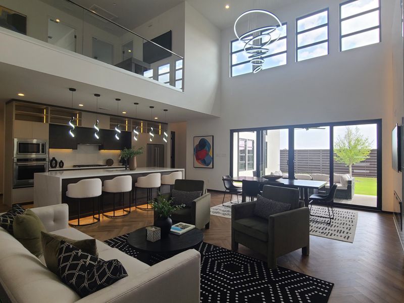 A modern living area with high ceilings, sleek kitchen, stylish lighting, and large windows opening to a patio.