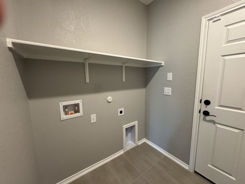A functional laundry room with light gray walls, tiled flooring, and a white shelf for storage.