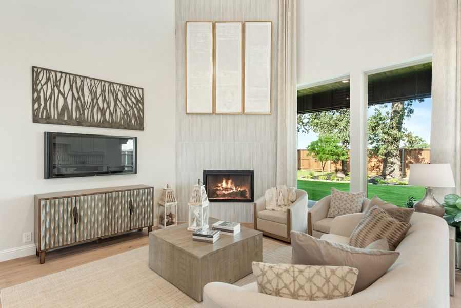Living room with high ceilings, lit fireplace, neutral seating, wall-mounted TV, and large windows overlooking a backyard Living room with high ceilings, lit fireplace, neutral seating, wall-mounted TV, and large windows overlooking a backyard
