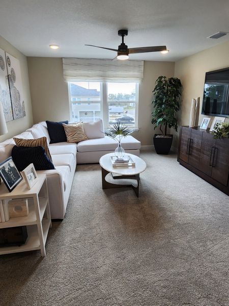 A cozy living room featuring a plush sectional, modern fan, large TV, and natural light through wide windows.